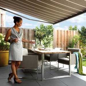 Eastbourne Shutters & Awnings - a photo of a woman in a light grey knee length cowl neck sleeveless dress. She isin a back garden patio of a home. She is placing a carafe of water onto an outdoor dining table which is set with four chairs. There are two throws over the arm of one chair, one is blue and one is green. There is an awning extended over head and lots of foliage visible in the picture.