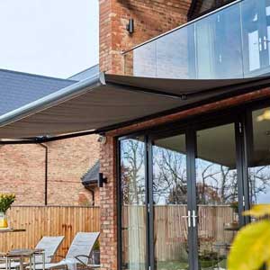  Trusted Shutter & Awning Installation in East Sussex - a photo of a grey awning extended at the back of two story home. The top story has a glass balcony and below that the awning is extended over double glass doors. There are lounge style outdoor chairs and a small outdoor side table as well.