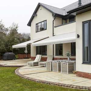  East Sussex Shutter & Awning Company - a photo of two awnings extended above windows at the back of a home. The back patio is semi circular in shape, leading onto lawn. It has two lounge style chairs as well as an outdoor dining table and chairs. There is also a covered barbecue.