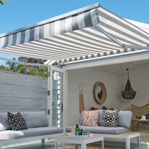 Local Shutter & Awning Company - a photo of a black and white striped awning extended from the front of a summer house or garden room with indoor and outdoor seating in grey, and wicker decorations in the summer house
