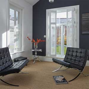 Full Height Shutters - a photo of a modern style living room or relaxing space. There are two leather and chrome sofas, These are a modern style with no arms and curved chrome legs. There are two large full height windows which are shuttered with full height shutters. These are white. There is a chrome table in the corner with a vase with orange flowers in it.