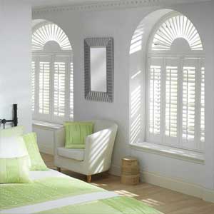 Special Shape Shutters - custom fit rounded top white shutters in a bedroom, the furniture is white with bright lime green cushions and throw accessories