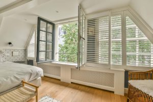 65ca41e17375e35a564f0bf0_comfortable-bed-light-bedroom faux wood shutter blinds fitted into a shaped bedroom window - you can see a bed, wicker chair, the bedroom has a wooden floor and a radiator which has a cover under the window