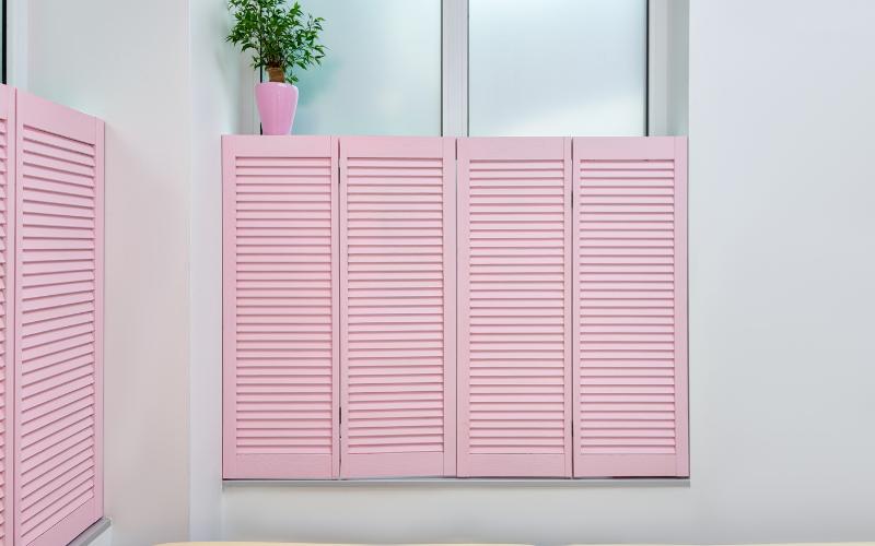 WhatsApp Image 2025-08-01 at 14.31.13 pink cafe style solid wood shutters fitted in a window with a small plant in a pink vase.
