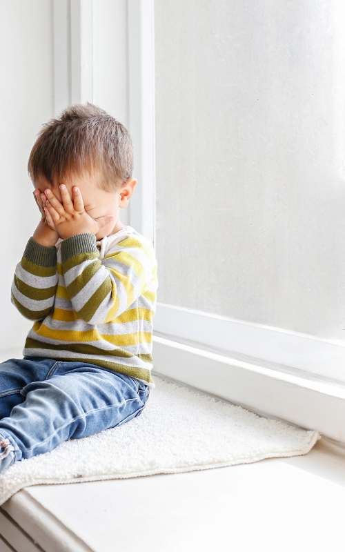 vertical website images (500 x 800 px) (1) child safe shutters - a small child sits on a windowsill - the window has neither blinds, curtains nor shutters. the child has their hands over their face as though playing peekaboo; they are wearing a striped jumper and blue jeans